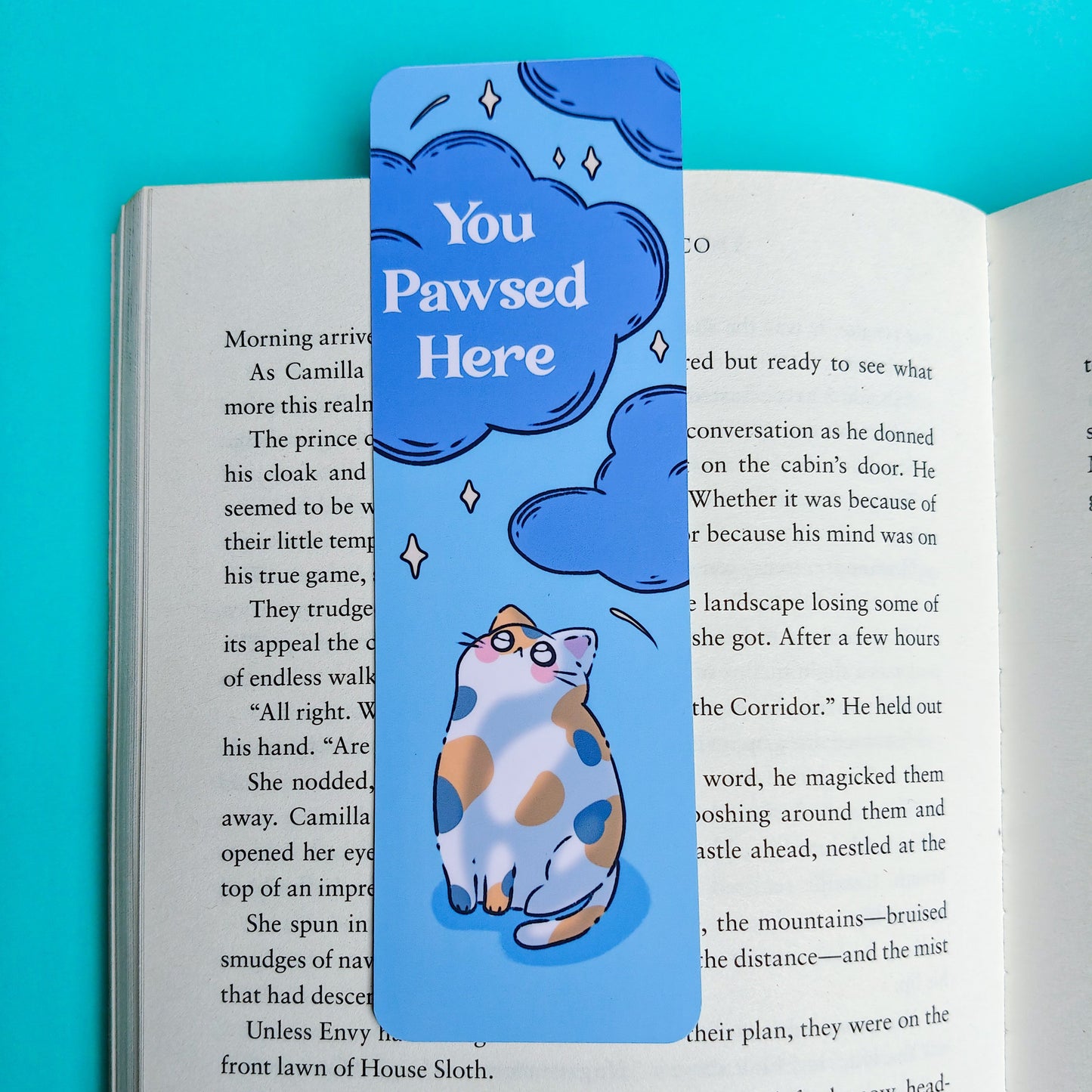 A printed book page with black text and a colorful illustration of a cat wearing glasses, with a blue speech bubble reading 'You Pawsed Here,' shown against a bright turquoise background.