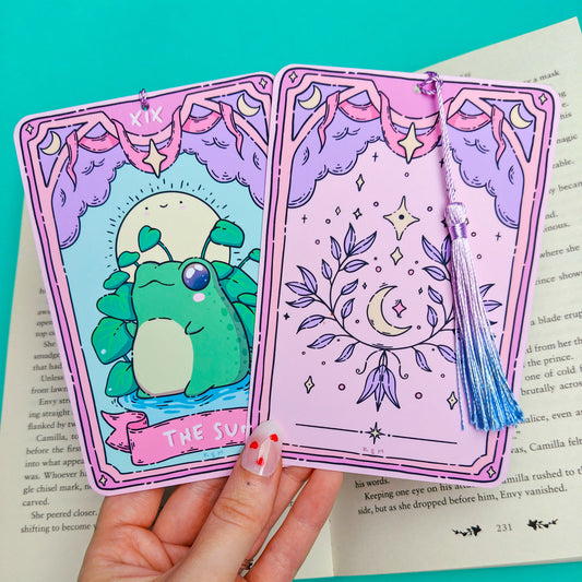 A hand holds two decorative tarot-style cards in front of an open book on a bright turquoise background. The left card is titled "The Sun" and shows a green frog under a yellow sun; the right card is lavender with a crescent moon, stars, and a blue tassel.