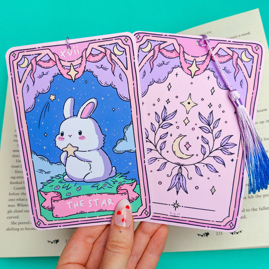 A hand holds two illustrated tarot-style cards with a blue tassel bookmark over an open book, against a bright turquoise background. The left card shows a white bunny in a starry blue scene labeled 'THE STAR,' and the right card has a pink celestial design with a crescent moon and stars.