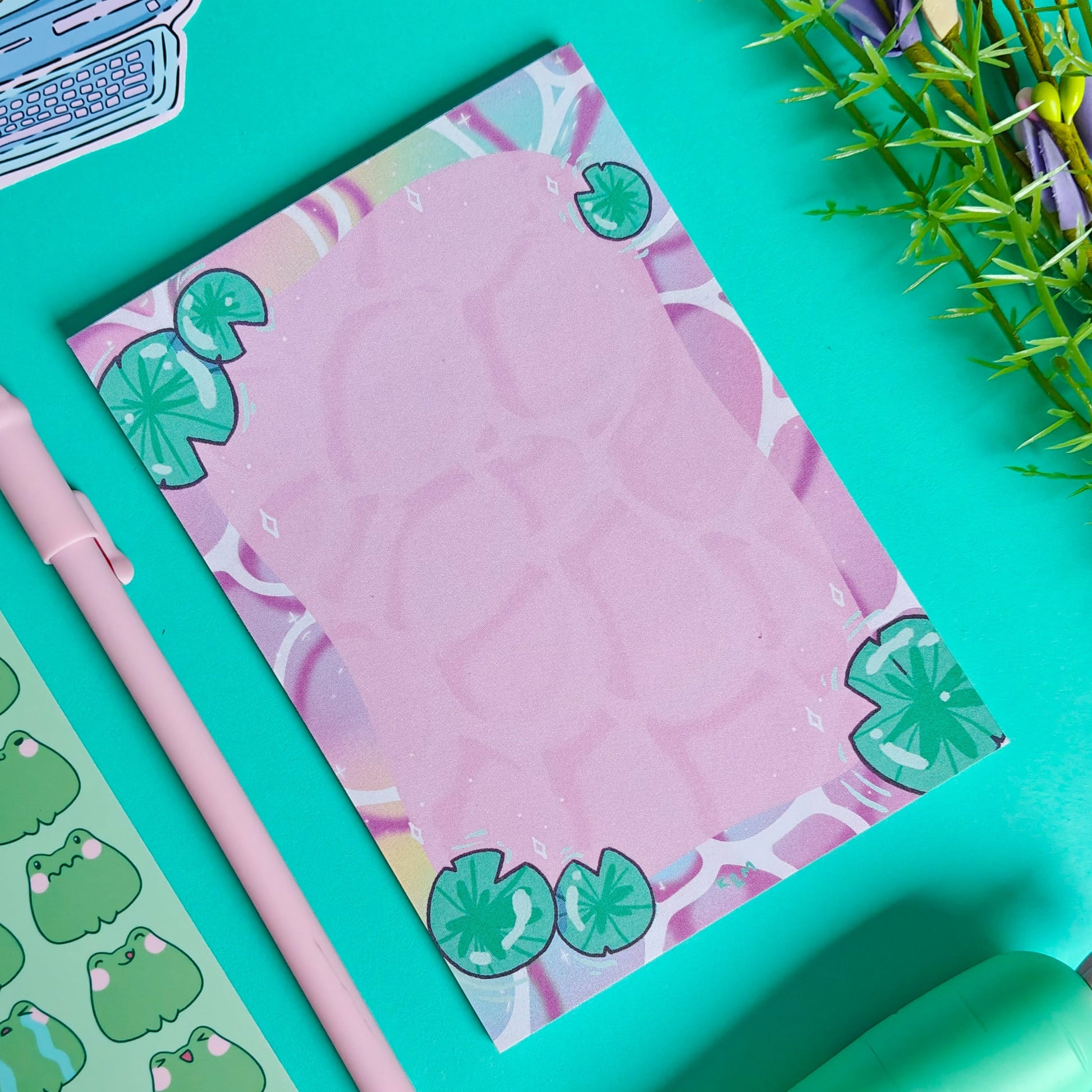 Notebook with green leaf design, pink pen, and green stickers on a teal background