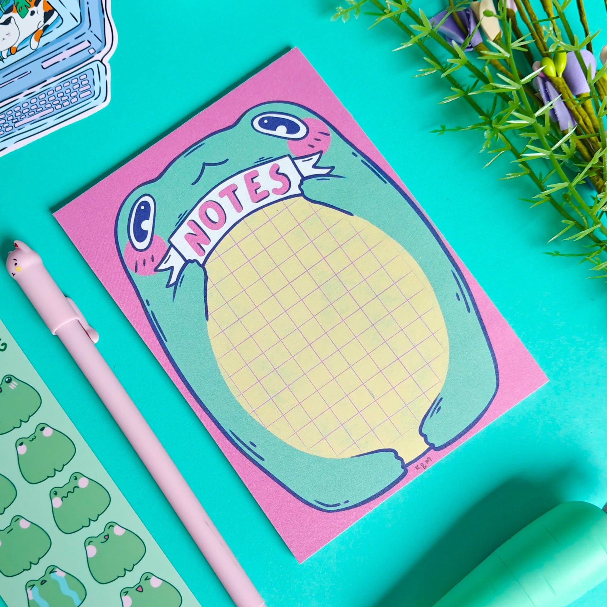 Notepad with frog design on a teal background with stationery items.