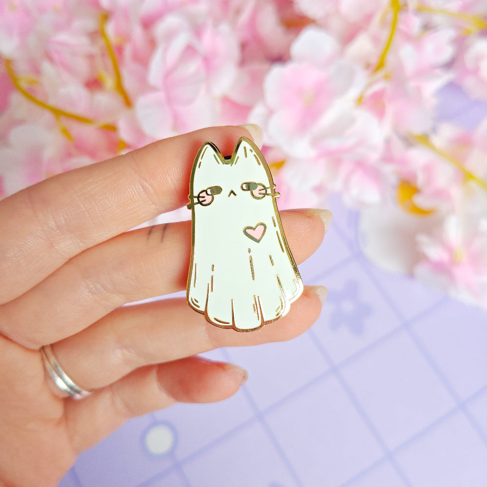 Hand holding a gold cat-shaped pin with a heart on a pink and purple floral background