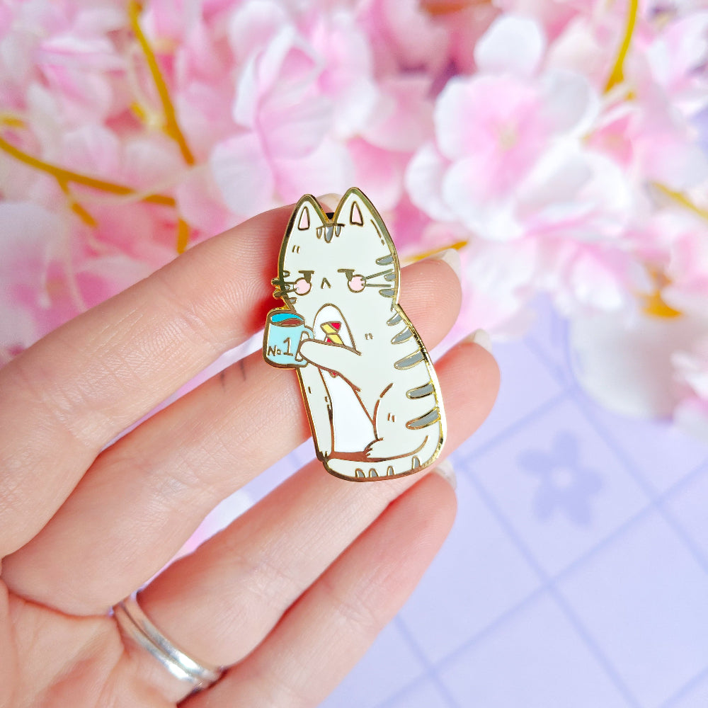 Cat-themed enamel pin held by a hand with a soft, blurred floral background