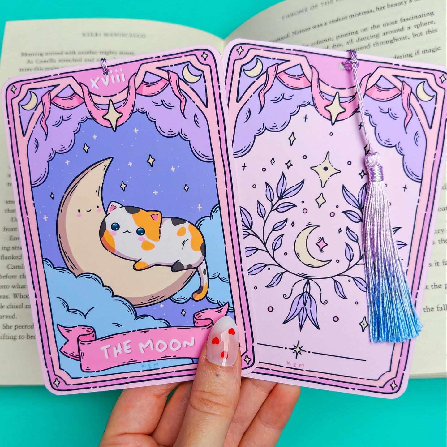 A hand holds two illustrated tarot-style cards with purple borders and celestial artwork, including a cat on a crescent moon and a blue tassel, in front of an open book on a bright turquoise background.