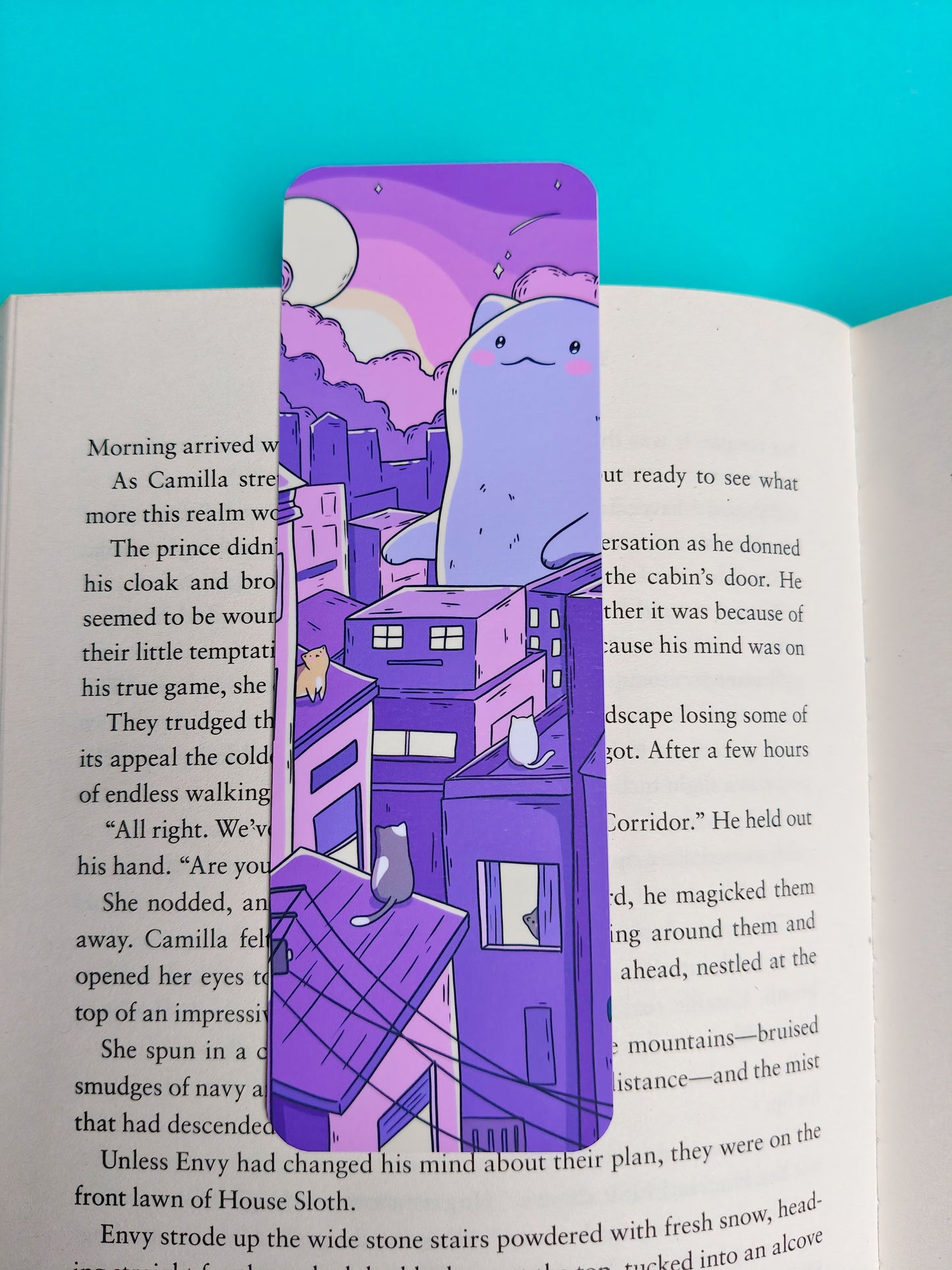 A purple illustrated book bookmark featuring a cartoon character and castle, placed on an open book page against a bright turquoise background.