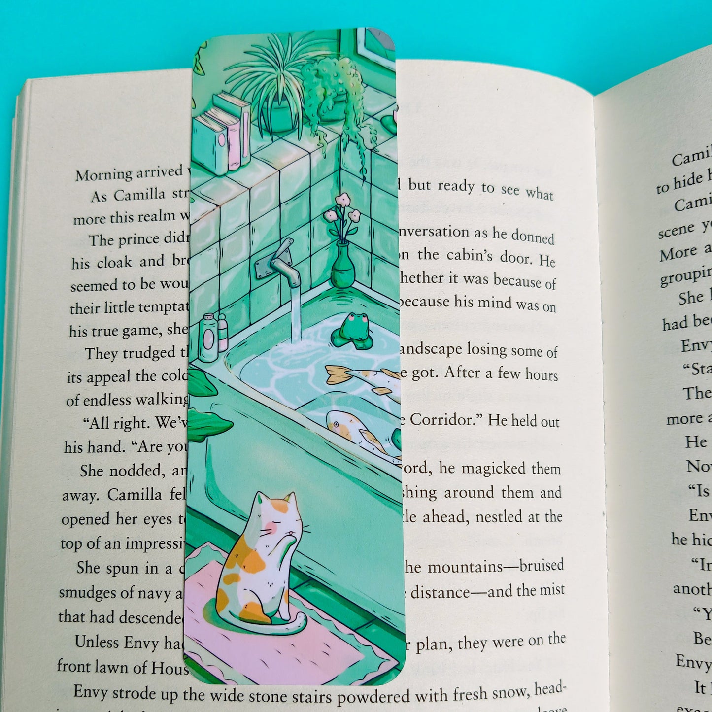An open book spread showing printed text and a colorful illustrated page insert featuring a whimsical green dragon-like creature in a fantasy landscape, photographed against a bright turquoise background.