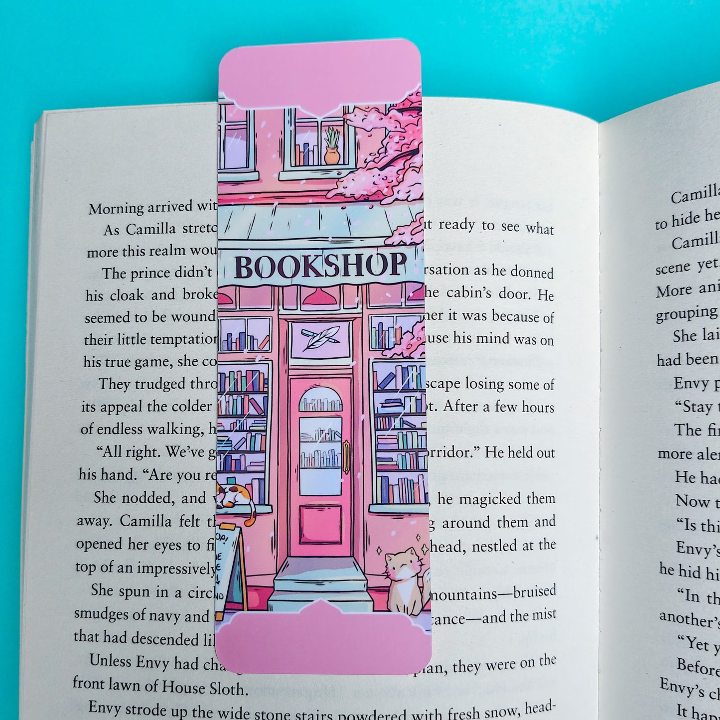 A pink and purple bookmark shaped like a bookshop with a pink door and illustrated shelves, placed on an open book page against a bright blue background.